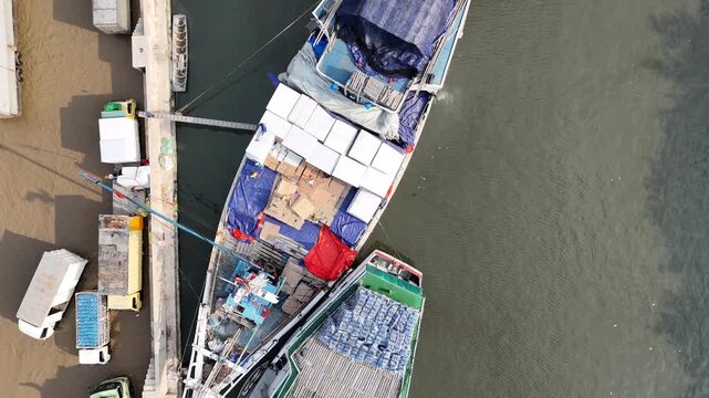 Top down aerial view of traditional wooden Pinisi cargo ships being loaded with white sacks at Sunda Kelapa port, featuring logistics trucks and a crane on the pier in Jakarta, Indonesia.