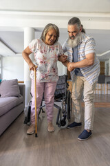 Fototapeta premium Senior woman and man assisting, woman floral blouse using cane while rising from wheelchair at home