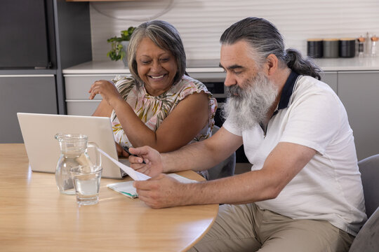 Senior couple sitting at round wooden table in kitchen, reviewing laptop and scattered documents