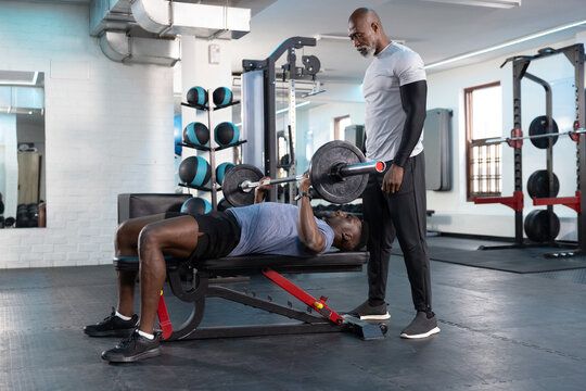 African American men bench pressing on flat bench in weight room while spotter assisting barbell