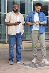 African American men standing on paved plaza holding smartphone and checking wristwatch near bench © wavebreak3