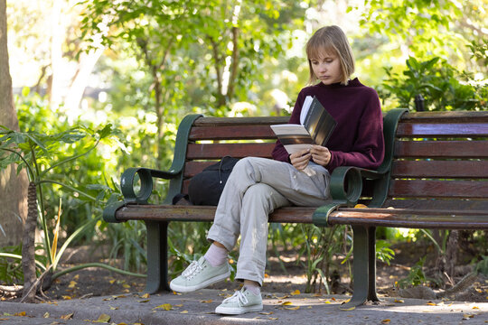 Woman sitting on wooden park bench reading paperback book with black backpack and maroon sweater