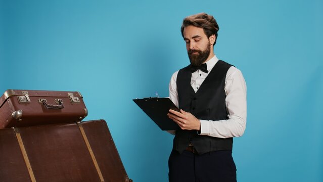 Doorman verifying checklist of guests, holding register log on clipboard to check hotel visitors in studio. Employee checking reservations on official ledger for archive, taking notes.