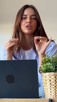 Confident young businesswoman speaking and gesturing during a video conference call from her modern office, actively engaging with her audience while sitting at her desk with a laptop