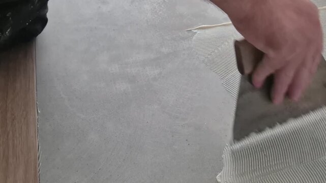 Close-up shot of a worker applying adhesive glue onto a prepared floor surface with a notched trowel before installing parquet flooring.