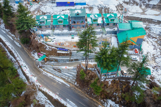Sonamarg, India - 05 February 2025: Aerial view of modern houses with green roofs on a snowy hillside surrounded by pine trees and a winding mountain road.