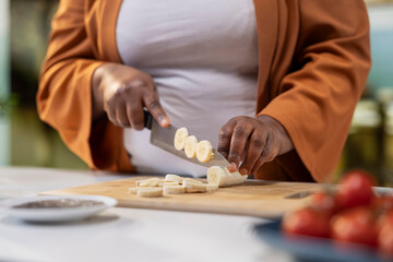 Obraz premium Close up of black woman vlogger chopping a banana on cutting board, adding slices of fruits on top of chia pudding, Filming easy recipe meal tutorial for healthy breakfast choice.