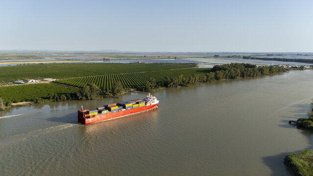 Coria del Rio, Spain - 02 June 2026: Aerial view of a large red container ship navigating the Guadalquivir river past lush green agricultural fields and rows of trees under a clear blue sky.