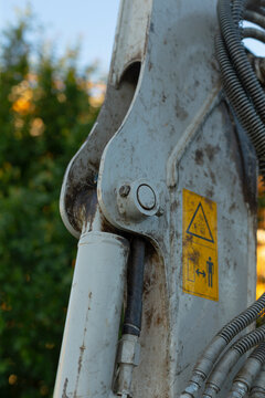 Detailed close-up of a heavy construction machine arm showing a worn metal joint, hydraulic cylinder, flexible hoses, and a yellow safety warning label