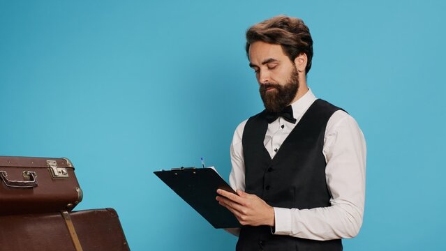 Bellboy reading checklist of guests, holding ledger or register on clipboard to verify list of travellers arriving at hotel. Staff checking reservations in official catalogue to keep evidence.