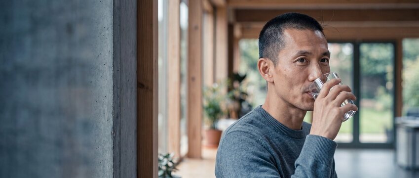 Asian man drinking water glass in modern home environment for hydration and wellness, standing by a concrete pillar near bright windows with houseplants in a relaxed morning routine lifestyle pictures
