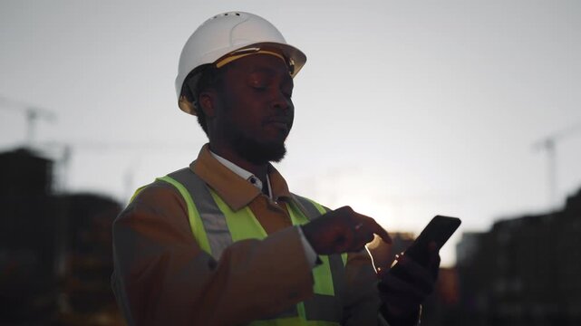 African builder chatting by mobile app in smartphone in construction site. Portrait of foreign professional foreman or engineer standing before building under construction, african american ethnicity
