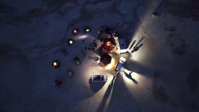 Aerial view of an illuminated campsite at night with tents and vehicles casting long light beams across the arid desert landscape in Kazakhstan.