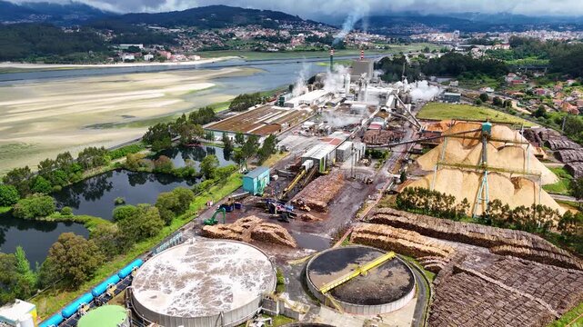Aerial view of the Biofabrica de Ence pulp mill with industrial chimneys, wood chip piles and water treatment tanks in Pontevedra, Pontevedra, Spain.