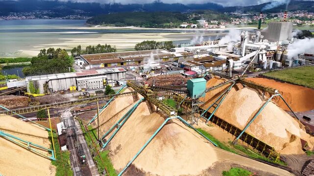 Aerial view of Biofabrica de Ence pulp mill featuring large wood chip piles, conveyor belts, and factory buildings near the coast in Pontevedra, Pontevedra, Spain.
