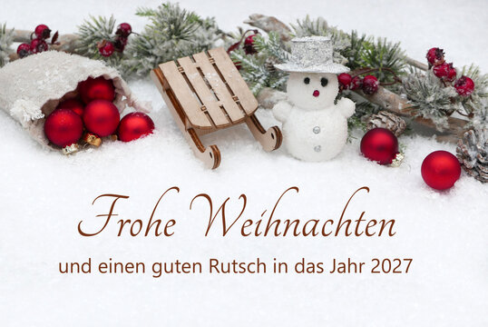 Weihnachtsgru&szlig;karte im Schnee mit kleinen Schneemann, einem Holzschlitten und roten Christbaumkugeln. Im Vordergrund steht der Schriftzug &bdquo;Frohe Weihnachten und einen guten Rutsch in das Jahr 2027