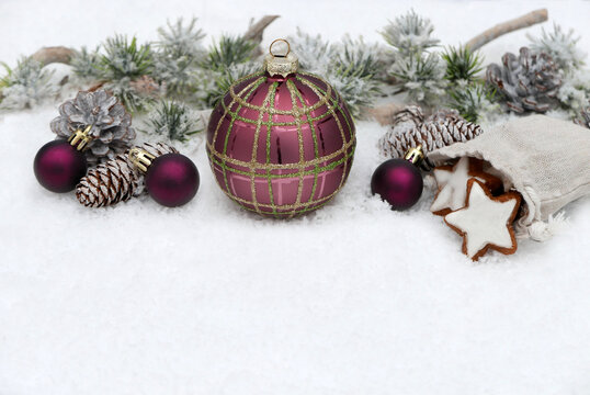 Weihnachtsdekoration mit viel Textfreiraum im Schnee. Eine gro&szlig;e Christbaumkugel mit kleinere Kugeln, Tannenzapfen, beschneiten Tannenzweige und ein Jutes&auml;ckchen mit Zimtsternen. 