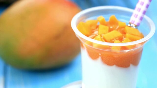 Fresh mango yogurt parfait in plastic cup, creamy tropical fruit dessert with orange mango chunks, healthy breakfast snack on blue background