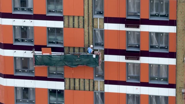 Construction worker in hard hat installing vinyl siding on high-rise building facade from aerial lift, summer exterior finishing with safety harness