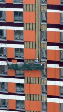 Construction worker in hard hat installing vinyl siding on high-rise building facade from aerial lift, summer exterior finishing with safety harness
