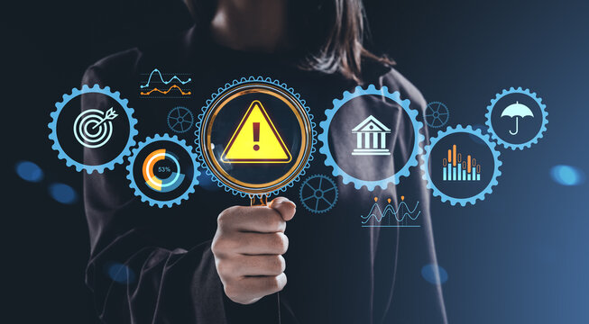 Risk analysis concept with hand holding magnifying glass highlighting warning icon among digital business symbols on dark background.