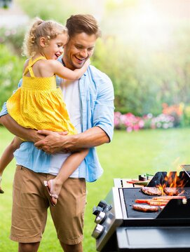 A joyful father holds his daughter while grilling meat on a barbecue in a sunny backyard. Family bonding and outdoor cooking.