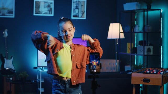 Joyful kid dancing in dimly lit home studio interior, producing content for online channel with smartphone on tripod. Smiling child doing viral dance choreography in living room