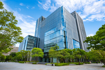 Obraz premium Modern high-rise office building with glass facade under a partly cloudy sky. Lush green trees surround the base