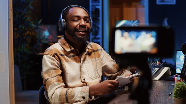Man recording interview with professional camera in studio, reviewing notes to ask pertinent questions to celebrity. Journalist filming with equipment, wearing headphones and talking on mic with guest