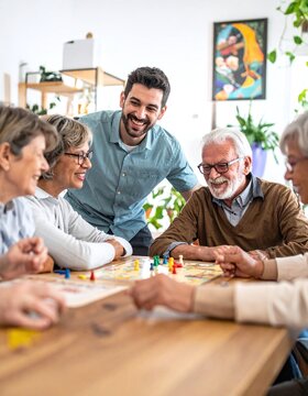 A cheerful group of senior friends enjoys playing a board game with a young male caregiver, showcasing themes of active aging, community support, elder care, and social bonding.