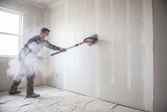 Construction worker sanding drywall in a room, creating dust clouds and preparing for finishing work.