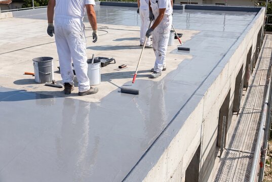 Construction workers in white overalls applying waterproofing sealant to a flat rooftop with rollers.