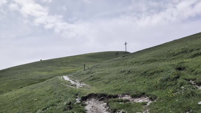 Pendio erboso con sentiero che sale verso una grande croce sulla cima della collina, sotto un cielo parzialmente nuvoloso. Atmosfera naturale, tranquilla e luminosa