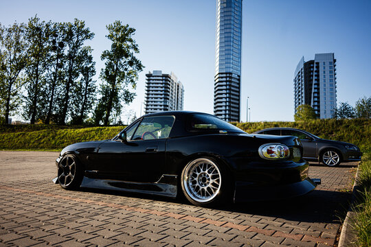 Low black Mazda MX5 sports car parked on a paved lot in Katowice Poland with urban skyline reflections aftermarket wheels and lowered stance