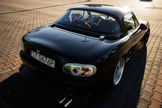 Shiny black Mazda sports car parked on a sunlit brick parking lot in Katowice, Poland with lowered stance and custom wheels at golden hour