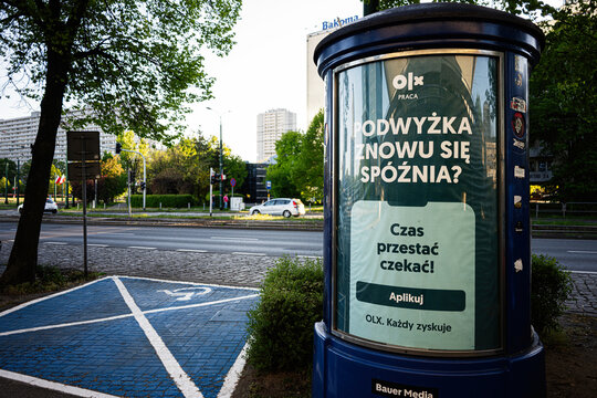 Street advertisement for OLX job offers on a cylindrical billboard in Katowice, Poland by an urban sidewalk and parking space