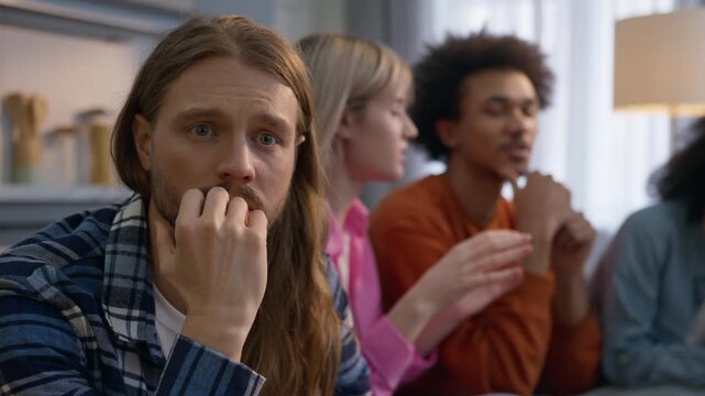 Despair anxious Caucasian man sitting on couch at home frustrated male thinking problem ignoring friends while multiracial African American women men speaking together social stress loneliness emotion