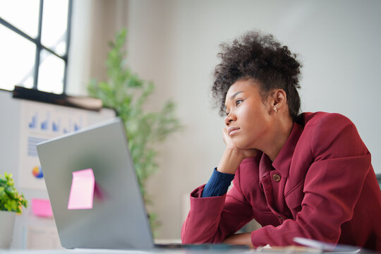 Black businesswoman feeling overwhelmed, experiencing burnout and fatigue at work, looking away from laptop while procrastinating on a boring task in a modern office