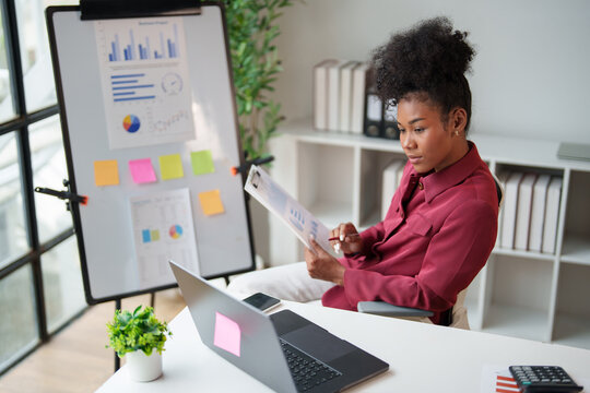 African American businesswoman analyzing financial data, reviewing statistics and business reports on documents and a laptop in a modern office for smart planning and development