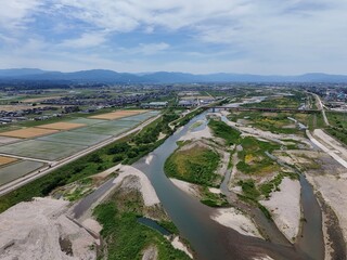 Fototapeta premium Wide river curves through farmland and towns. Sky holds scattered clouds above rolling hills. Riverbanks show patches of green and exposed earth. Distant structures blend with open fields