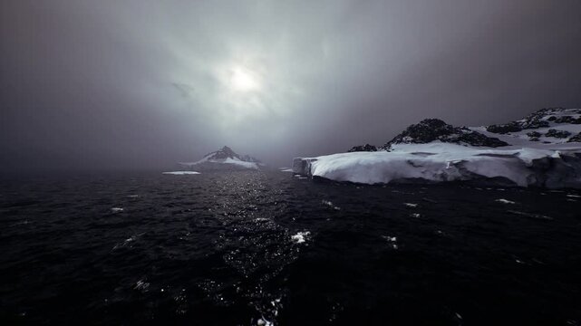 Distant icy peak beyond dark sea under moody sky, rugged ridge rising from glacier, cold water in foreground with scattered floes, sense of remote wilderness