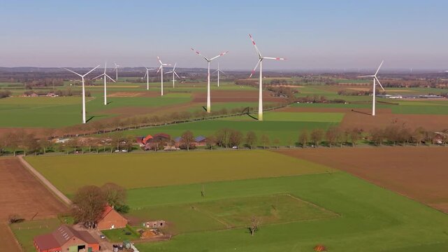 An aerial drone view of a vast rural landscape featuring scattered farmsteads and cultivated fields with multiple large wind turbines lining the horizon under a clear sky.