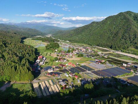 Lush mountains frame a quiet village nestled in green valleys. Rows of paddies and fields stretch below a bright blue sky. Sunlight bathes rooftops and trees in golden warmth. Village life hums gently
