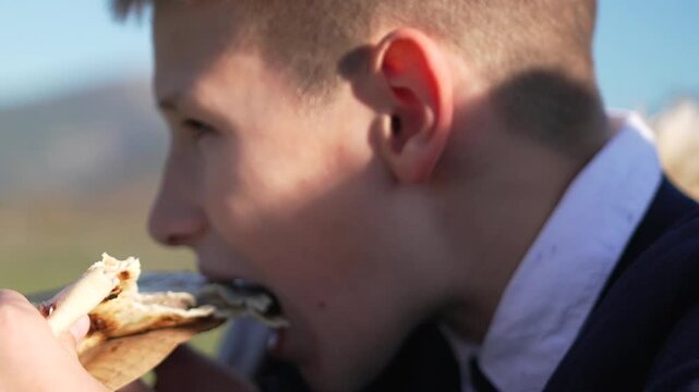 Boy eating pita bread outdoors on sunny day, enjoying a simple picnic lunch with mountain scenery.