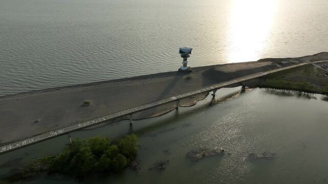 Horizontal drone aerial footage frame capturing the long wooden pedestrian bridge in Anaklia, showcasing the river delta meeting the Black Sea beach, a small green island, and coastal resort