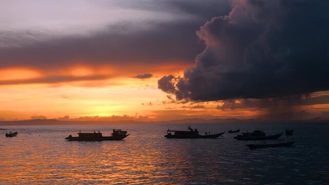 SILHOUETTE: A line of anchored boats in glowing water while a towering storm cloud spills rain over the horizon. Mix of tropical calm with incoming stormy weather at sunset in the Mentawai Islands.