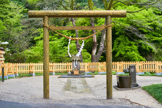 A traditional wooden torii gate stands at the entrance to a Japanese shrine, featuring a sacred shimenawa rope with white zigzag-shaped shide papers hanging across the top beam. The weathered wooden