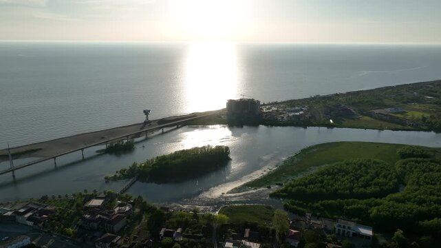Horizontal drone aerial footage frame capturing the long wooden pedestrian bridge in Anaklia, showcasing the river delta meeting the Black Sea beach, a small green island, and coastal resort