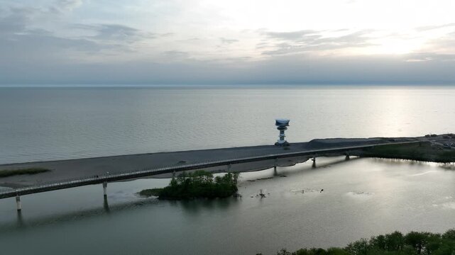 Horizontal drone aerial footage frame capturing the long wooden pedestrian bridge in Anaklia, showcasing the river delta meeting the Black Sea beach, a small green island, and coastal resort