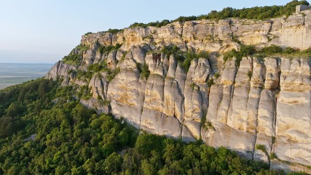 High vertical limestone rocks cliff face texture green forest trees growing mountain base sun light summer landscape aerial drone view Madara Bulgaria